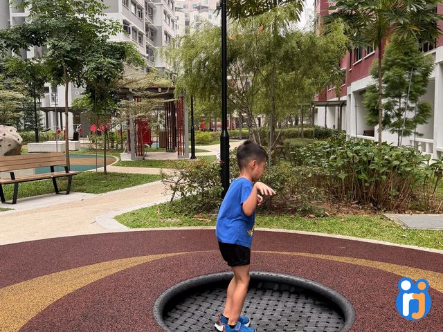 A boy bounces on a round trampoline set into the ground, on a maroon and yellow rubber floor.