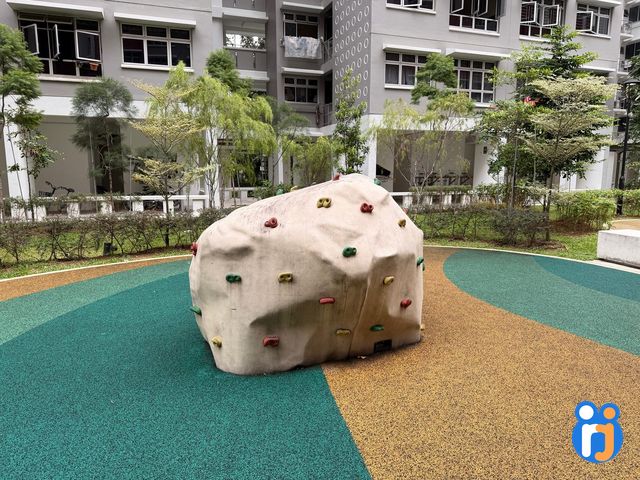 A pale climbing boulder dotted with colourful holds, on a green and yellow rubber floor in front of HDB blocks.