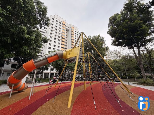 A tall yellow-framed rope pyramid with an orange tube slide, on a red, maroon and ochre rubber floor near HDB blocks.