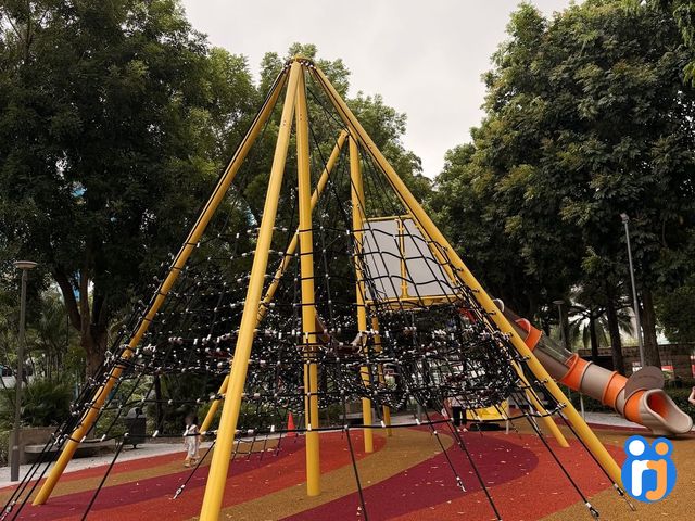 Looking up at the tallest rope pyramid, its yellow poles meeting above a web of black climbing ropes, with an orange tube slide beside it.