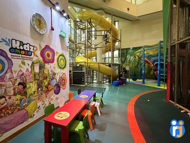 The play hall, with a painted mural wall and small bright tables in front of the yellow tube-slide tower.