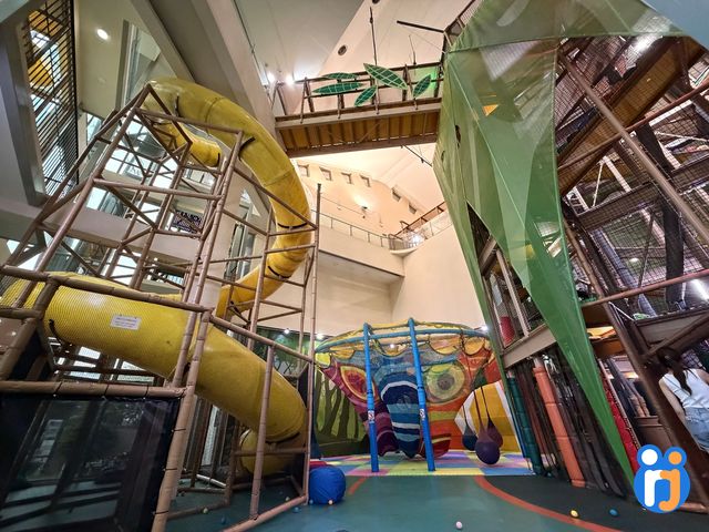 Yellow tube slides wind down the tall timber-and-net climbing structure towards the rainbow crochet net.